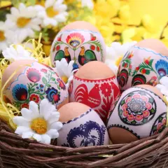 Koszyk z wielkanocnymi pisankami ozdobionymi tradycyjnymi wzorami kwiatowymi i kogutami, otoczony sztucznymi kwiatami.