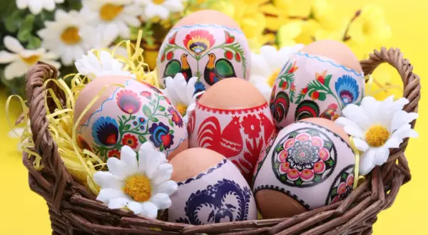 Koszyk z wielkanocnymi pisankami ozdobionymi tradycyjnymi wzorami kwiatowymi i kogutami, otoczony sztucznymi kwiatami.